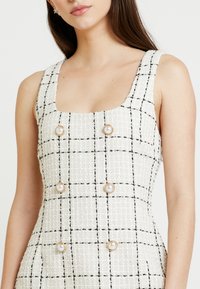 Vit tweedklänning med svart rutmönster, fyrkantig halsringning och breda axelband. Har pärlliknande knappar med gulddetaljer.