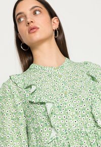 Vestido floral verde com detalhes em babados e frente com botões, apresentando flores brancas e pretas sobre um fundo texturizado.