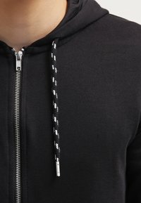 Schwarze Zip-Hoodie mit gerippter Struktur, ausgestattet mit einem silbernen Reißverschluss und schwarz-weißen geflochtenen Kordeln. Einfaches und funktionales Design.