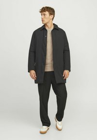 Manteau noir au design simple, avec un col et une fermeture à bouton. Porté sur un pull beige et associé à un pantalon noir et des baskets.