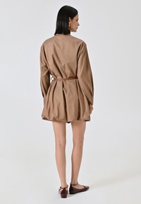 Bruine, textuur jurk met lange mouwen, gecinchde taille met een riem en een geplooide rok. Gemaakt in combinatie met bruine platte schoenen. Neutrale achtergrond.