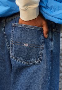 Siniset farkut, joissa on tasku, jossa näkyy pieni monivärinen logo. Käsi lepää takataskussa. Pehmeä kangas, jossa näkyvät saumat.