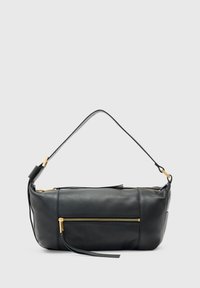 Sac en cuir noir porté à l'épaule avec des fermetures éclair dorées, une poche avant et une poignée fine, présenté sur un fond gris clair.