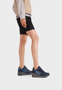 Sneakers blu navy con una combinazione di texture liscia e scamosciata, dotati di una suola con zeppa e di una cerniera laterale per un facile accesso. Pantaloni corti neri e una camicia a maniche lunghe a righe.