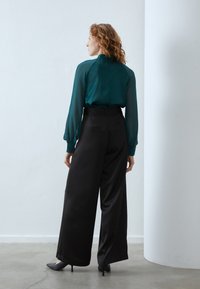 Blouse en voile teal à manches longues associée à un pantalon large taille haute noir, présentant une texture lisse et un design minimaliste.