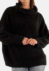 Eine Frau trägt einen schwarzen strukturierten Rollkragenpullover mit langen Ärmeln, die Hände an der Taille gefaltet, und hellblaue Jeans.
