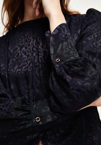Marineblaue Bluse mit subtilen Leopardenmuster; besitzt luftige Ärmel und goldfarbene Knopfakzente an den Bündchen.
