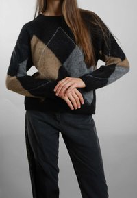 Persona che indossa un maglione a scacchi nei colori nero, grigio e beige e pantaloni scuri, in posa con le mani delicatamente intrecciate davanti.