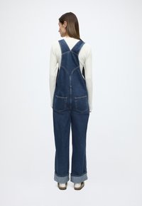 Tumeda sinise värvusega teksariidest püksid lõdva lõikega, millel on kaks tagataskut, reguleeritavad rihmad ja ülespööratud ääred. Materjalil on sile tekstuur.