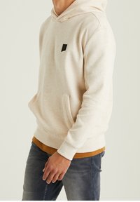 Sweat à capuche beige clair en tissu texturé, avec une poche kangourou à l'avant et un petit logo noir sur la poitrine. Porté par-dessus une chemise foncée.
