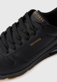 Black leather sneaker with lace-up design, featuring a textured surface, gold 'SKECHERS' branding, and rubber sole.