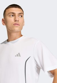 Witte sportieve T-shirt met korte mouwen, voorzien van een grijs logo en een strakke grijze designelement langs de schouder. Zachte textuur.