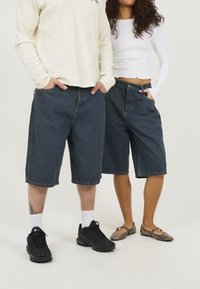 Pantalones cortos de mezclilla en azul oscuro con un corte holgado, a la altura de la rodilla, con bolsillos frontales y costuras visibles; combinados con calzado en color contrastante.