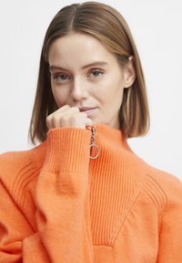Pull-over orange en demi-zip avec texture côtelée, doté d'une tirette de zip ronde et d'un design côtelé diagonal sur les épaules. Matériau doux en mélange de laine.