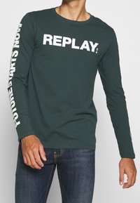 Man wearing dark green long-sleeve shirt with "REPLAY" on chest and "FUTURES STARTS NOW" text on left sleeve, paired with blue jeans.