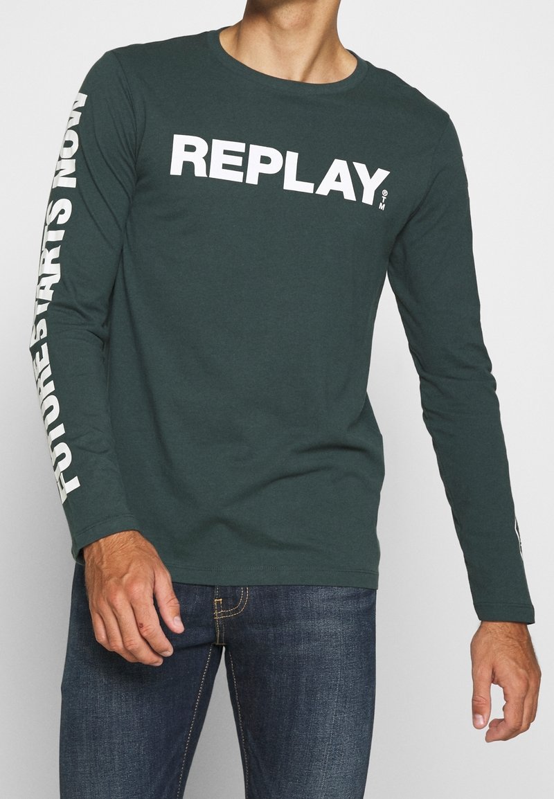 Man wearing dark green long-sleeve shirt with "REPLAY" on chest and "FUTURES STARTS NOW" text on left sleeve, paired with blue jeans.