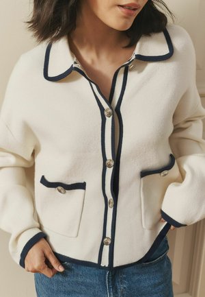 Woman wearing a cream cardigan with navy trim and silver buttons, paired with blue jeans, standing indoors with hands near pockets.