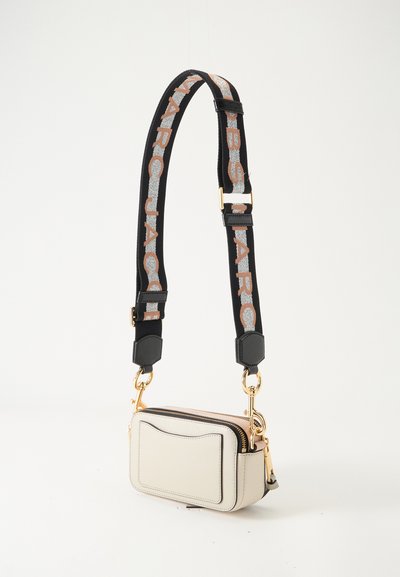 Μικρό λευκό τσαντάκι crossbody με μπροστινή τσέπη, χρυσά εξαρτήματα και φαρδιά μαύρη ρυθμιζόμενη λουρά με μεγάλο μπεζ κείμενο.