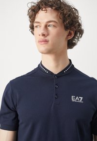 Navy polo shirt made of cotton, featuring a button-up collar, short sleeves, and a textured collar with square accents and a logo.