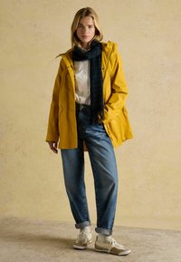 Geltona vandeniui atspari striukė su kapišonu, dėvima ant baltos marškinių. Mėlyni aukštu liemeniu džinsai ir kreminės spalvos sportiniai bateliai su baltais akcentais. Tamsiai margas šalikas.