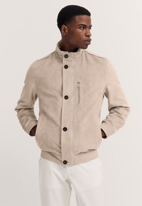 Beige suede jacket with a stand collar, black buttons, and a zippered chest pocket. Soft texture, fitted design, and side pockets included.