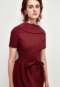 Burgundské šaty s krátkými rukávy, velkým límcem a uvázaným páskem v pase. Tkanina má hladkou texturu a strukturovaný design.