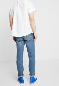 Camicetta bianca a maniche corte con orlo arrotondato, abbinata a jeans in denim azzurro. Sneakers blu con accenti bianchi completano l'outfit.
