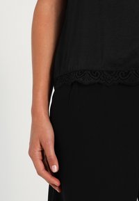 Schwarzes Oberteil mit einem Spitzenbesatz am Saum und einer glatten Textur. Eine Hand befindet sich neben dem Saum und zeigt eine neutrale Nagellackfarbe.