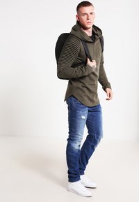 Felpa verde oliva con design delle maniche a costine, abbinata a jeans blu consumati e sneakers bianche. Zaino nero appeso a una spalla.