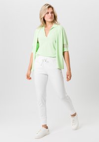 Hellgrünes Poloshirt mit kurzen Ärmeln und V-Ausschnitt, kombiniert mit weißen Hose mit Kordelzug. Das Model trägt weiße Sneakers. Schlichtes, sportliches Design.