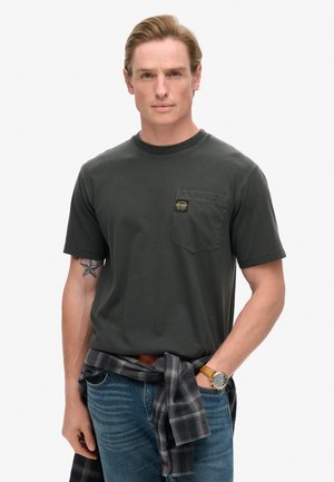 T-shirt di cotone verde scuro con taschino sul petto, logo dorato, su uno sfondo semplice. Abbinato a jeans blu e una camicia a quadri nera annodata in vita.