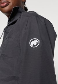 Giacca nera con superficie texture, dotata di un logo bianco di un elefante ricamato sul maniche sinistro. Chiusura a zip, colletto alto.