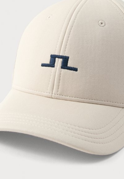 Ανοιχτό μπεζ καπέλο μπέιζμπολ με ένα μικρό, σκούρο μπλε κεντητό αφηρημένο σύμβολο που βρίσκεται στο κέντρο της μπροστινής πλευράς.