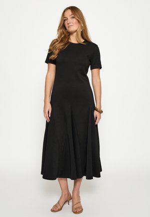 Ženska z dolgimi lasmi, oblečena v črno midi obleko s kratkimi rokavi, rjavim zapestnim trakom in gojzarji z naramnico, stoji pred enobarvnim ozadjem.