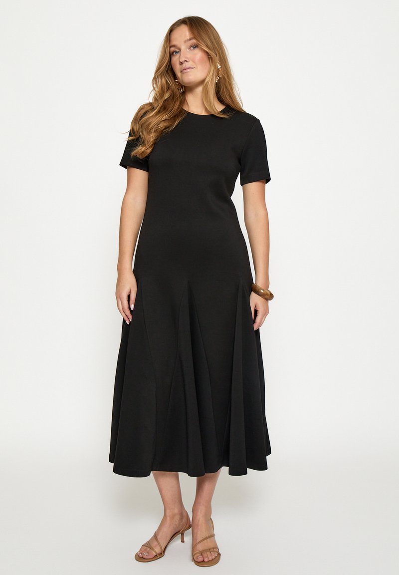 Frau mit langen Haaren, die ein schwarzes Midi-Kleid mit kurzen Ärmeln, ein braunes Armband und nude-farbene Riemchenabsätze trägt, steht vor einem einfarbigen Hintergrund.