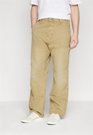 Mężczyzna noszący luźne beżowe spodnie typu cargo i białe trampki, stojący z rękami w kieszeniach na jednolitym białym tle.