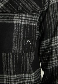 Tissu à carreaux noir et gris avec un bouton noir et une petite pièce noire présentant un motif abstrait blanc sur une poche de vêtement.
