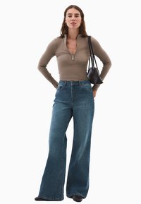 Ung kvinde iført brun langærmet zip-up top, højtaljede brede blå jeans, sort håndtaske over skulderen, stående med hænderne på hoften.