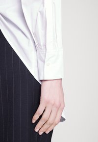 Poignet de chemise en coton blanc avec un bouton, associé à un vêtement rayé noir. Les manches retroussées révèlent une texture lisse et des lignes nettes.