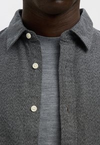 Mann trägt eine graue, geknöpfte Wollhemdjacke über einem hellgrauen, gerippten Strickpullover, zeigt Kragen und oberen Brustbereich.