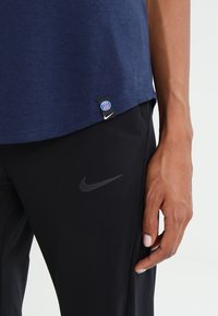 Närbild av en person som bär en marinblå skjorta med Paris Saint-Germain- och Nike-logotyper, samt svarta byxor med en grå Nike-swoosh.