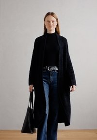 Dlhý, voľný čierny cardigan s textúrovaným úpletom, nosený na čiernom roláku, doplnený o riflové nohavice s vysokým pásom a čiernu taštičku.
