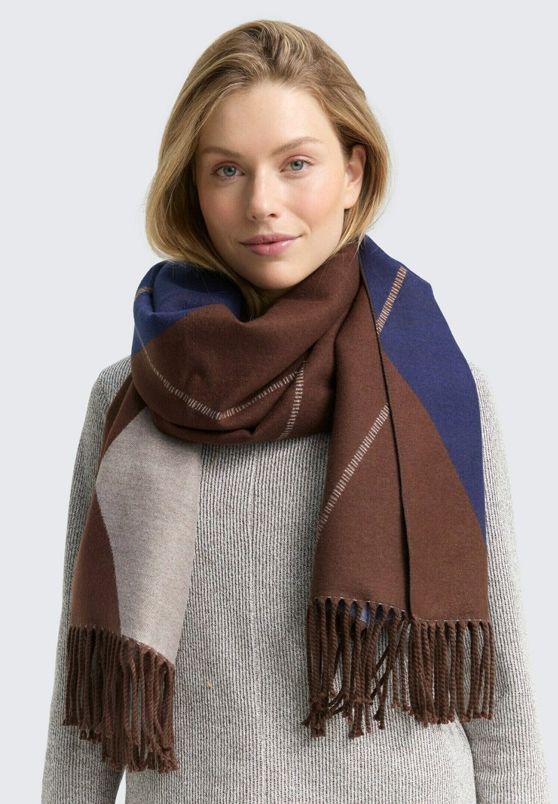 Bruine, marineblauwe en beige sjaal met een patroon en franjes, gemaakt van zacht materiaal. Draagt om de nek gewikkeld tegen een gestructureerde trui.