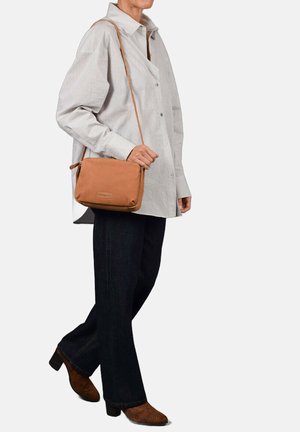 Sac en cuir marron en bandoulière avec une texture lisse, de forme rectangulaire et un logo discret, accompagné d'une chemise à rayures et d'un jean foncé.