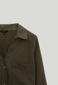 Chemise à manches longues vert olive foncé avec col, boutons à l'avant et poche visible sur le côté gauche de la poitrine, sur un fond clair uni.