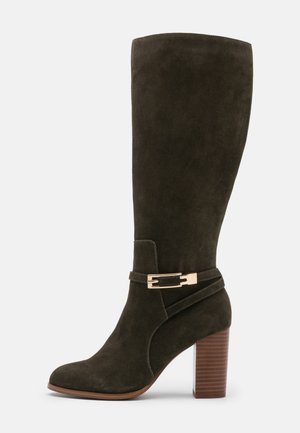 Ciemnozielony zamszowy kozak za kolano na obcasie z drewnianych bloczków, z paskiem z klamrą w kolorze złota wokół kostki.