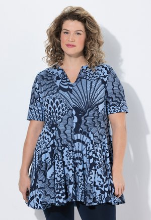 Kvinde med krøllet hår iført en blå og marine mønstret kortærmet tunika, stående mod en ensartet lys baggrund.