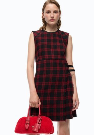 Femme portant une robe sans manches à carreaux rouges et noirs tenant un sac à main rouge texturé avec des poignées en cuir et une petite étiquette.