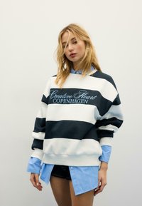 Felpa a righe blu navy e bianca con la scritta "Creative Heart COPENHAGEN", abbinata a una camicia azzurra con bottoni e pantaloncini di jeans.