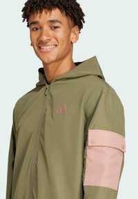 Green hooded jacket with a zip closure, featuring a pink accent on the arm and a small logo on the chest. Smooth fabric texture.